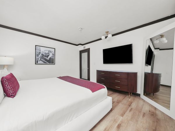 A bright hotel-style bedroom with a large white bed, purple accent pillows, wooden dresser, wall-mounted TV, a full-length mirror, and light wood flooring.