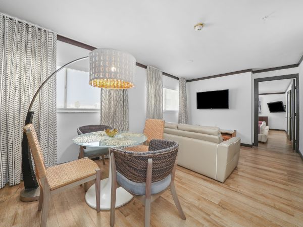 A modern, open-plan living and dining area with light wood floors, a round glass dining table, wicker chairs, a beige sofa, and a wall-mounted TV.