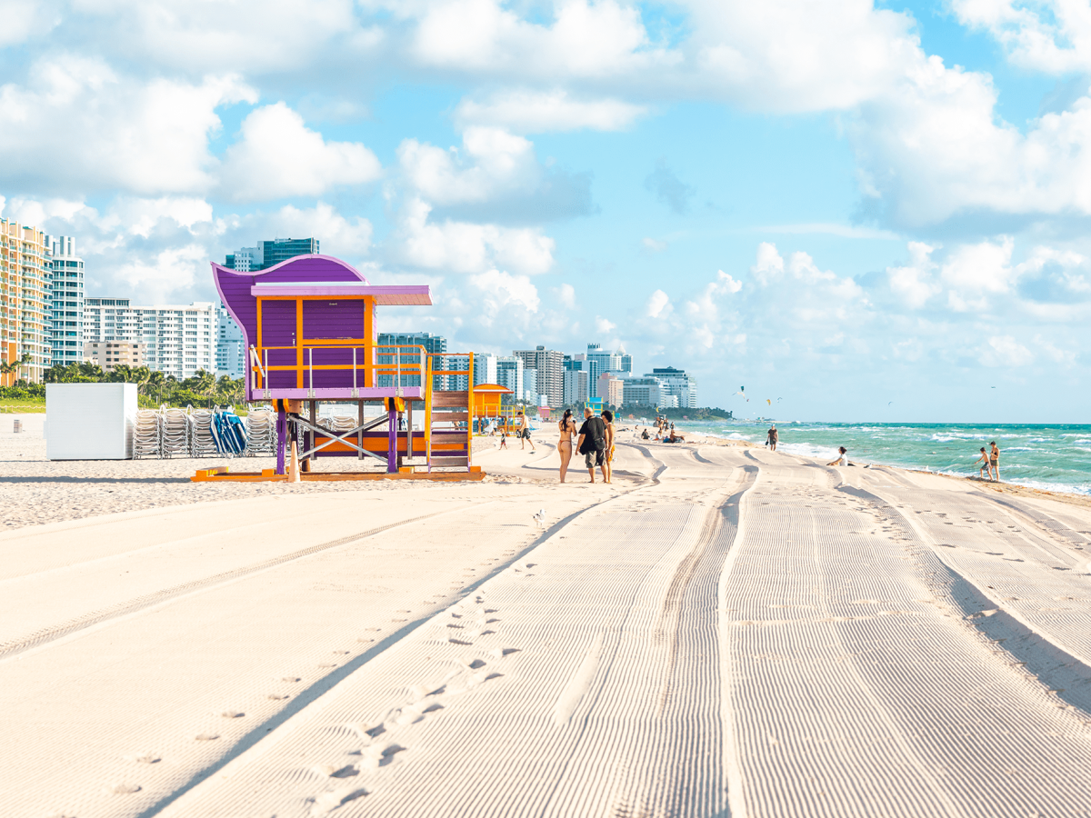 A sunny beach with white sand, a colorful lifeguard hut, high-rise hotels in background, and turquoise ocean under a blue sky with fluffy clouds.