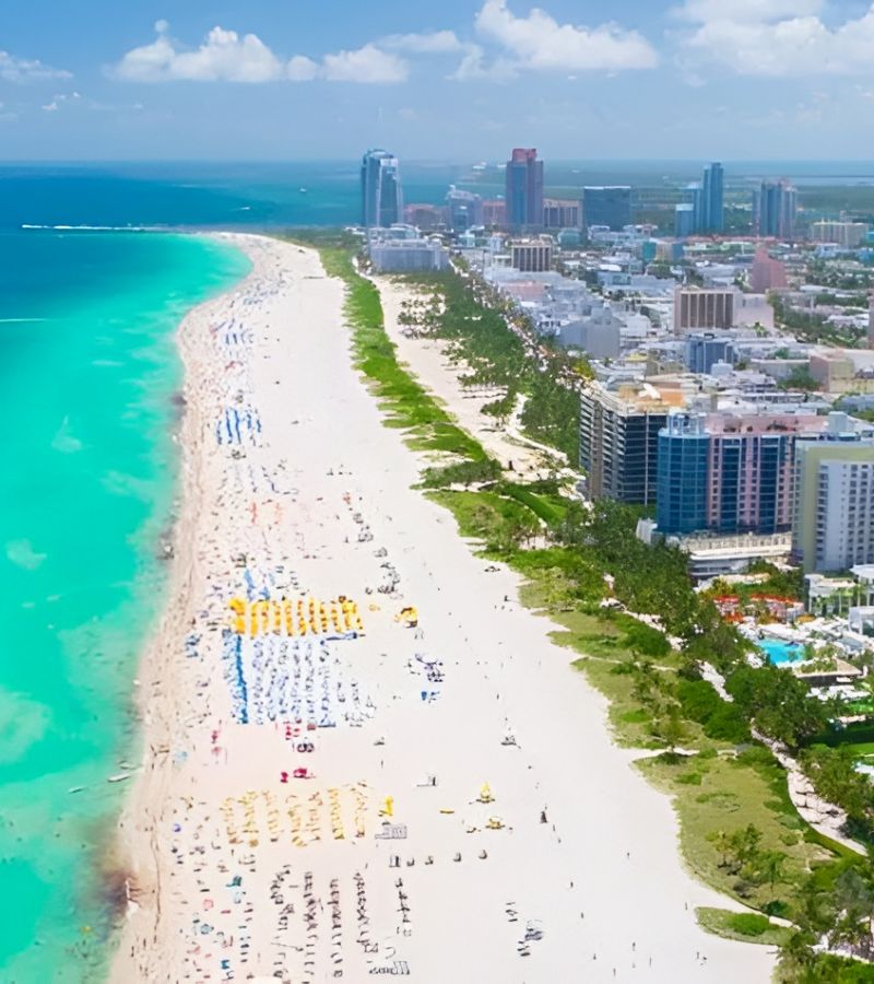 An aerial view of a beach with white sand and turquoise water, lined with buildings and trees, and a cityscape in the background.