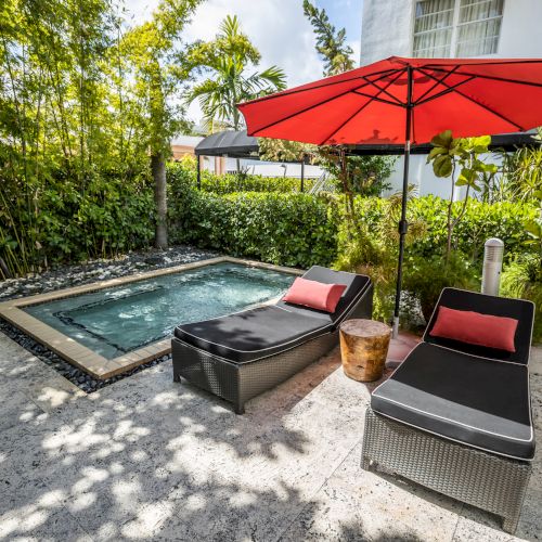 Tropical backyard with a small pool, red patio umbrella, and lounge chairs surrounded by lush greenery.