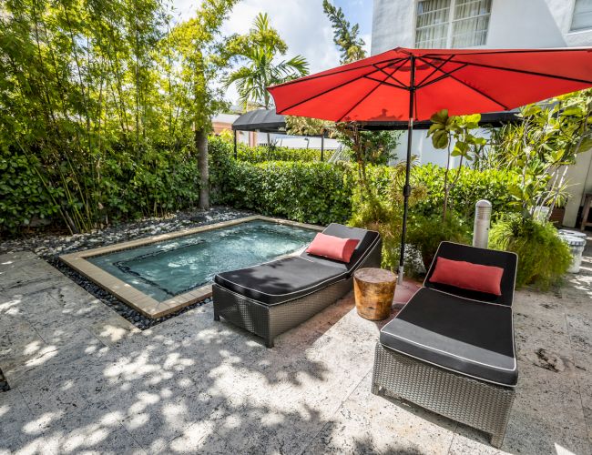Tropical backyard with a small pool, red patio umbrella, and lounge chairs surrounded by lush greenery.