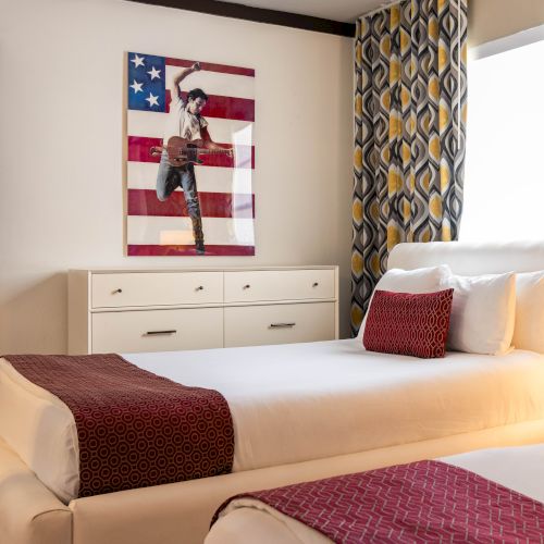 A bright bedroom with a made bed, dresser, patterned curtains, and a wall artwork of a guitarist against the U.S. flag.