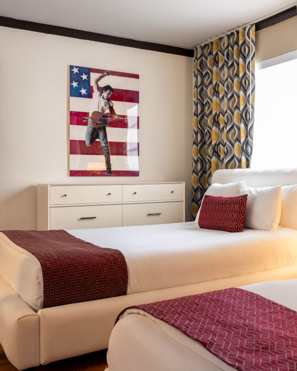 A bright bedroom with a made bed, dresser, patterned curtains, and a wall artwork of a guitarist against the U.S. flag.