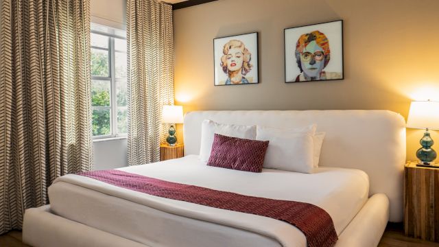 A neatly made bed with white linens and a red throw blanket, flanked by two nightstands with lamps, in a room with a window and patterned curtains.
