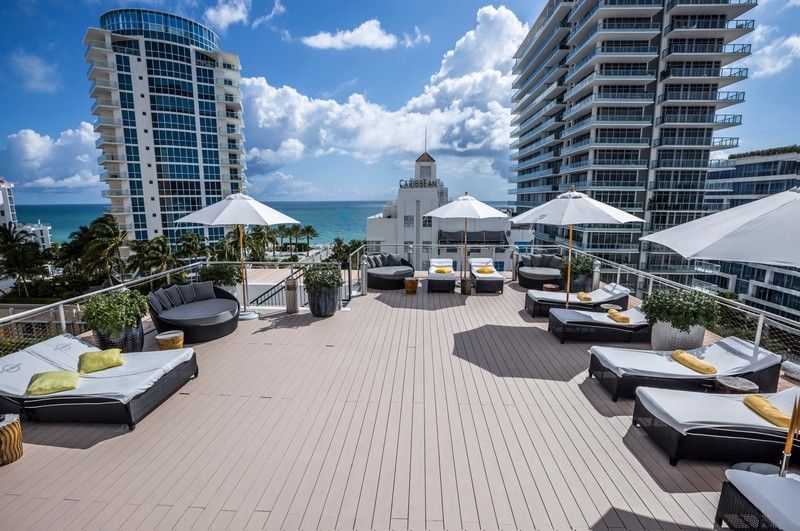 A rooftop pool area with lounge chairs, umbrellas, and a view of the ocean and city skyline.